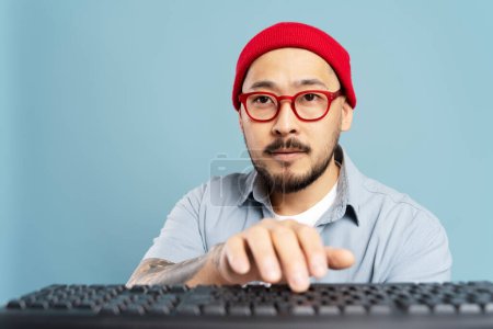 Foto de Retrato de una pensativa redactora asiática escribiendo en el teclado, trabajando proyecto freelance, comunicación en línea. Elegante hombre coreano, diseñador con sombrero hipster, gafas rojas aisladas sobre fondo azul - Imagen libre de derechos
