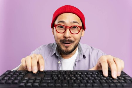 Téléchargez les photos : Hipster coréen excité jouant à un jeu vidéo. Incroyable homme asiatique portant chapeau rouge, lunettes shopping en ligne isolé sur fond violet. Portrait de pigiste émotionnel, rédacteur sur clavier - en image libre de droit