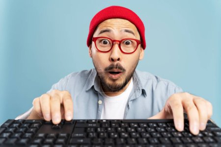 Foto de Asombroso hombre asiático con sombrero rojo, anteojos escribiendo en el teclado, compras en línea aislado sobre fondo azul. Emocional juego hipster coreano. Retrato del proyecto de trabajo del programador emocional - Imagen libre de derechos