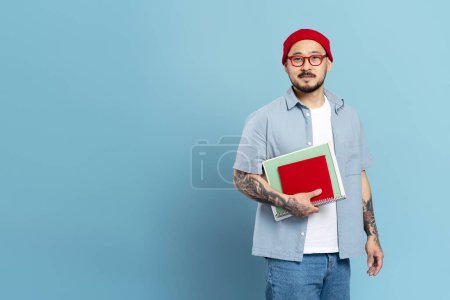 Foto de Sonriente estudiante coreano guapo sosteniendo libros aislados sobre fondo azul, concepto de educación. Retrato de hombre asiático freelancer con tatuaje elegante, usando sombrero rojo, gafas, espacio de copia - Imagen libre de derechos