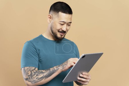 Handsome smiling asian man holding digital tablet watching videos, communication online isolated on beige background. Happy Korean hipster shopping online, ordering food on website. Technology concept