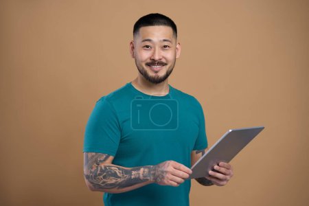 Portrait of confident smiling asian student holding modern digital tablet, isolated on beige background. Happy Korean hipster with e book in hand looking at camera. Technology concept