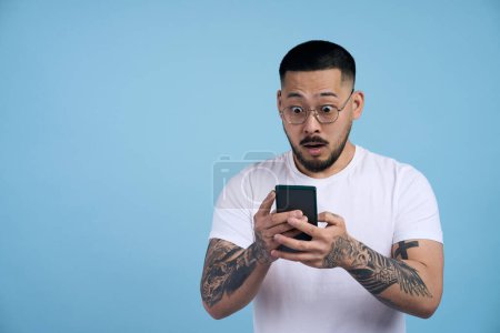 Foto de Retrato del hombre asiático conmocionado sosteniendo el teléfono móvil leyendo noticias de última hora aisladas sobre fondo azul. Resultado del examen de lectura de estudiantes emocionales. Concepto tecnológico - Imagen libre de derechos