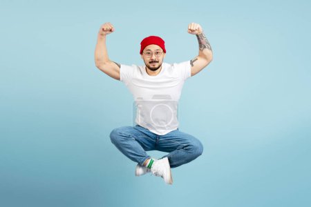 Photo for Strong asian man, bodybuilder with stylish tattoo showing muscles isolated on blue background. Muscular Korean male jumping high in lotus pose, looking at camera. Gym workout, sport, healthy lifestyle - Royalty Free Image