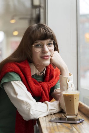 Photo for Portrait of beautiful young woman wearing glasses, stylish sweater, coffee break in modern cafe. Attractive female hipster using smartphone, looking at camera - Royalty Free Image