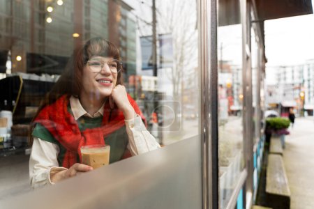 Foto de Hermosa mujer joven sonriente con gafas con estilo sosteniendo vidrio con bebida mirando a la ventana, espacio de copia. Mañana, concepto de pausa para café - Imagen libre de derechos