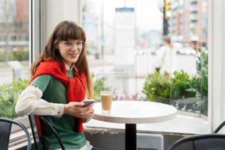 Foto de Retrato de mujer atractiva con gafas con estilo, sosteniendo el teléfono móvil, sentado en la cafetería, mirando a la cámara. Chica joven navegando, charlando - Imagen libre de derechos