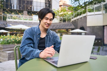 Téléchargez les photos : Portrait d'un homme asiatique confiant et souriant utilisant la communication par ordinateur portable, ayant un appel vidéo assis à l'extérieur. Programmeur japonais performant sur le lieu de travail. Concept technologique - en image libre de droit