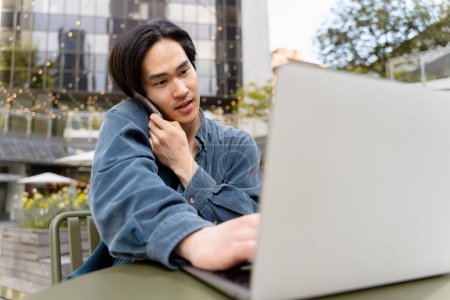 Photo for Young japanese businessman using laptop, talking on mobile phone, working online outdoors. Handsome asian man sitting at workplace. Remote job, technology concept - Royalty Free Image