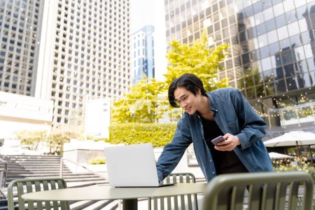 Photo for Smiling asian freelancer working on computer, using laptop, holding mobile phone, standing near workplace outdoors. Portrait of young japanese entrepreneur working in cafe on street. Business concept - Royalty Free Image