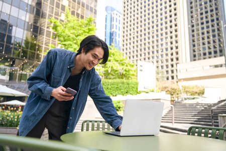 Téléchargez les photos : Heureux étudiant japonais vérifiant ordinateur portable e-mail et tenant téléphone mobile dans le quartier des affaires. Technologies et concept de communication - en image libre de droit