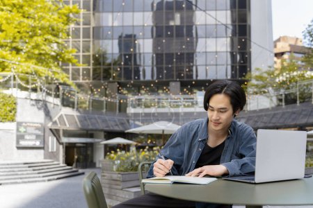 Téléchargez les photos : Portrait de jeune étudiant asiatique en ligne étudiant l'écriture dans un cahier assis sur le campus universitaire. Homme d'affaires japonais confiant travaillant à l'extérieur sur son lieu de travail. Concept d'éducation en ligne - en image libre de droit