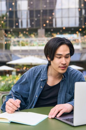 Téléchargez les photos : Homme asiatique, pigiste utilisant ordinateur portable prenant des notes, projet de travail à l'extérieur. Vue verticale de coûteux beau Japonais étudiant, apprendre des langues, concept d'éducation en ligne - en image libre de droit