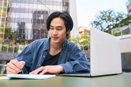 Foto de Hombre asiático, freelancer usando computadora portátil tomando notas, trabajando proyecto al aire libre. Estudiante japonés guapo pensativo que estudia, aprendiendo idiomas, concepto en línea de la educación - Imagen libre de derechos