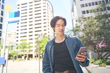 Foto de Joven sonriente hombre asiático sosteniendo teléfono inteligente usando aplicación móvil, lectura de mensajes de texto, comunicación en línea caminando por la calle urbana. Japonés hipster chico compras en línea al aire libre - Imagen libre de derechos