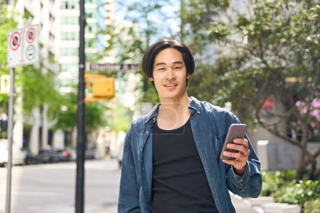 Photo for Young smiling Japanese man holding smartphone using mobile app, looking camera, communication online walking on urban street. Asian hipster guy shopping online outdoors - Royalty Free Image