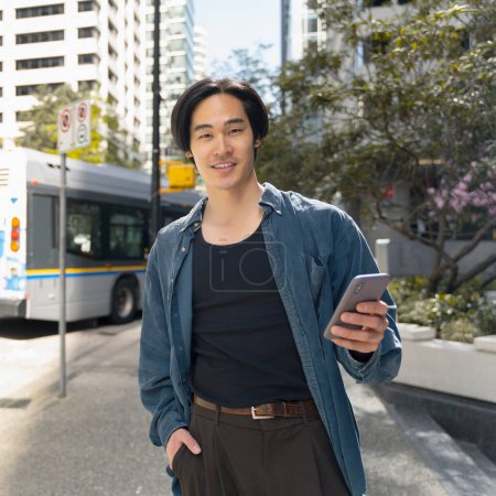 Téléchargez les photos : Portrait d'un bel homme asiatique tenant un téléphone portable, regardant la caméra à l'extérieur, dans la rue de la ville. Succès japonais homme d'affaires vérifier le courrier. Concept de communication en ligne, technologie moderne - en image libre de droit