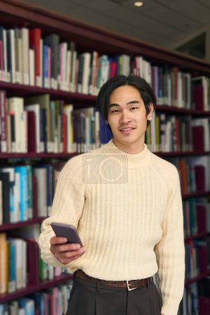 Photo for Vertical view of smart Japanese university student holding mobile phone in modern library, education concept. Handsome asian man in bookstore - Royalty Free Image