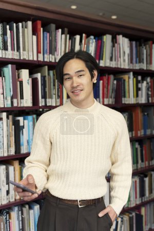 Foto de Sonriente hombre asiático de pie en la biblioteca universitaria, el uso de teléfonos inteligentes, el aprendizaje del idioma, la educación. Atractivo hombre japonés casual leyendo libros, mirando a la cámara. Concepto de aprendizaje en línea - Imagen libre de derechos