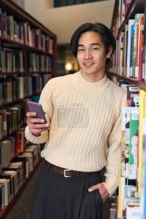 Téléchargez les photos : Vue verticale d'un étudiant japonais intelligent tenant un téléphone portable dans une bibliothèque moderne, concept éducatif. beau asiatique l'homme dans librairie - en image libre de droit