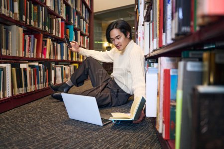 Téléchargez les photos : Japonais détendu étudiant ordinateur portable, tenant livre de papier et smartphone, se préparant à l'examen assis dans la bibliothèque universitaire, l'éducation. Asiatique étudiant apprentissage des langues - en image libre de droit