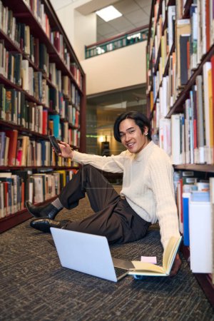 Photo for Vertical view of smiling Relaxed Japanese man studying laptop, holding paper book and smartphone, preparing to exam sitting in university library, education. Asian student looking at camera - Royalty Free Image