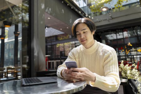 Photo for Stylish asian businessman holding mobile phone, sitting at workplace, using laptop. Modern japanese hipster checking mail, browsing. Technology concept, remote job - Royalty Free Image
