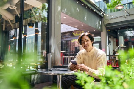 Téléchargez les photos : Jeune homme d'affaires japonais confiant utilisant le téléphone mobile, regardant la caméra et travaillant en ligne assis dans un café moderne. Beau sourire asiatique homme vérification email copie espace .Technology concept - en image libre de droit