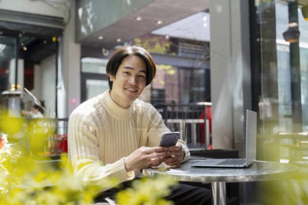 Téléchargez les photos : Un homme asiatique souriant tenant un téléphone portable, lisant un message texte regardant une caméra. Jeune homme d'affaires japonais confiant utilisant un ordinateur portable travaillant en ligne assis dans un café moderne. Concept technologique - en image libre de droit