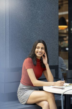 Foto de Retrato de una hermosa mujer india sonriente tomando notas en un cuaderno mirando hacia otro lado sentada en una cafetería moderna. Smart estudiante asiático feliz estudiando, aprendiendo el idioma sentado en el escritorio - Imagen libre de derechos