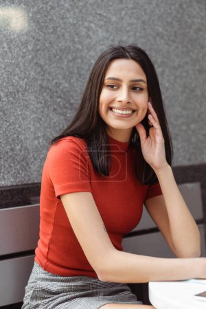 Photo for Vertical portrait of beautiful smiling Indian woman wearing stylish casual clothes looking away on the street. Cute asian student with brunette hair sitting in university campus. Education concept - Royalty Free Image