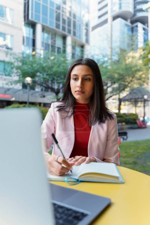 Photo for Serious smart asian student studying, learning language in university campus. Young pensive Indian businesswoman, freelancer using laptop, taking notes, planning project working online at workplace - Royalty Free Image
