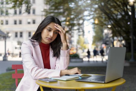 Téléchargez les photos : Stressé femme indienne fatiguée à l'aide d'un ordinateur portable de recherche d'informations, travaillant en ligne assis sur le lieu de travail. Étudiant asiatique sérieux étudiant, apprendre la langue, avoir mal à la tête. Concept d'éducation - en image libre de droit