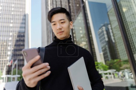 Téléchargez les photos : Attrayant asiatique étudiant bavarder sur téléphone mobile, tenant ordinateur portable à l'extérieur. Beau Chinois regardant dans le smartphone. Concept technologique - en image libre de droit