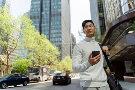 Foto de Atractivo turista asiático de pie en la calle de la ciudad sosteniendo el teléfono móvil mirando hacia otro lado. Hombre de negocios chino sonriente utilizando el teléfono inteligente al aire libre en la ciudad. Concepto de viajes, negocios - Imagen libre de derechos