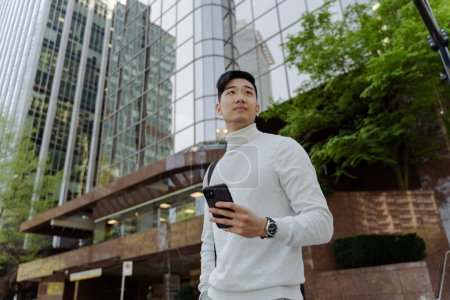 Téléchargez les photos : Portrait d'un étudiant asiatique élégant attrayant tenant un téléphone portable marchant en ville avec un sac détournant les yeux. Jeunes touristes chinois debout sur la rue de la ville urbaine. Concept de voyage - en image libre de droit