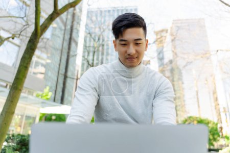Téléchargez les photos : Succès asiatique homme d'affaires en utilisant un ordinateur portable, travailler à l'extérieur, travailler en ligne. Attrayant pigiste chinois, programmeur professionnel travaillant à l'ordinateur. Concept technologique - en image libre de droit
