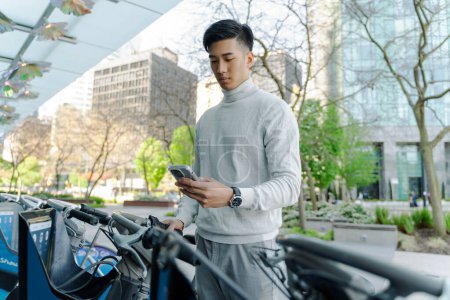 Téléchargez les photos : Homme asiatique sérieux tenant un téléphone mobile, scanner application mobile, location de vélos dans la rue. Attrayant touristique chinois acheter un service en ligne - en image libre de droit
