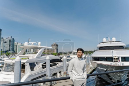 Téléchargez les photos : Portrait d'homme riche asiatique debout près de yachts chers, regardant loin à l'extérieur. attrayant chinois mâle posant pour photo, sur la rue - en image libre de droit