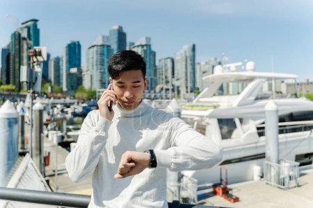 Photo for Confident asian man checking time, talking on mobile phone, standing near expensive yacht outdoors. Attractive chinese businessman in yacht club - Royalty Free Image