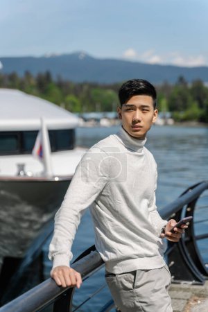 Téléchargez les photos : Portrait d'un homme d'affaires asiatique debout près d'un yacht, tenant son téléphone portable et regardant ailleurs. Homme chinois élégant voyageant sur yacht - en image libre de droit