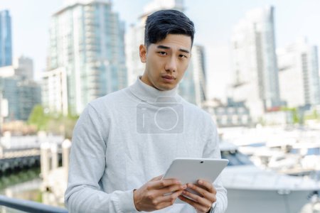 Téléchargez les photos : Pensif, sérieux homme d'affaires asiatique en utilisant une tablette numérique, fond debout de la ville urbaine. attrayant chinois homme regardant caméra - en image libre de droit