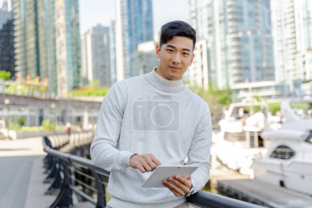 Foto de Exitoso hombre de negocios asiático utilizando tableta digital, de pie cerca del yate, mirando a la cámara. Hombre chino guapo en el club de yates moderno. Concepto empresarial - Imagen libre de derechos