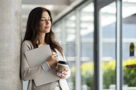 Photo for Tired beautiful woman with closed eyes holding laptop, drinking coffee in modern office, break. Smiling businesswoman working. Fashion model posing for picture, successful career - Royalty Free Image
