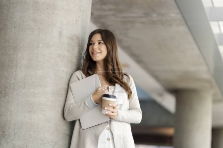 Photo for Smiling woman holding laptop and drink in paper cup looking away in modern office. Beautiful female during coffee break, working in office at workplace. Concept of business - Royalty Free Image