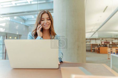 Téléchargez les photos : Portrait d'une femme souriante parlant sur un téléphone portable, utilisant un ordinateur portable, assise à l'espace de copie du lieu de travail. Beau projet confiant de planification de femme d'affaires, travaillant en ligne. Étudiante étudiante en bibliothèque - en image libre de droit