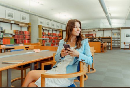 Photo for Beautiful, confident businesswoman holding mobile phone, checking mail, sitting at workplace, online shopping. Smiling female woman wearing blue jacket working in library, ordering, online shopping - Royalty Free Image