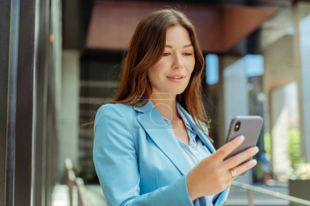 Beautiful smiling woman holding mobile phone, shopping online, reading text message, watching video in modern office. Successful freelancer receive payment. Technology, mobile banking concept