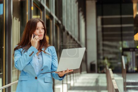 Téléchargez les photos : Portrait de belle femme d'affaires portant une veste bleue à l'aide d'un ordinateur portable et parlant sur un téléphone mobile à l'extérieur. Femme réussie travaillant en ligne, détournant les yeux, copier l'espace. Concept multitâche - en image libre de droit