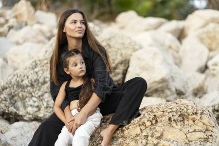 Photo for Beautiful pensive young mother with little daughter hugging, sitting on beach looking away. Happy family together on vacation, travel concept - Royalty Free Image